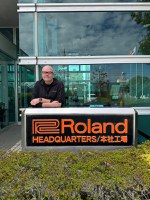 Paul McCabe in front of a glass-walled building, leaning on a sign with the words Roland Headquarters followed by Japanese characters in orange with black background. 
