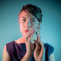 Erin Gee Erin Gee headshot in front of a teal-blue background, hands curved and nearly touching, vertically just below her chin, lightly grasping a silver rod or flute head joint.