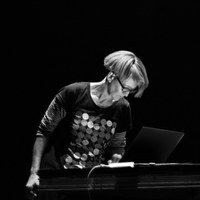 Monique Jean wearing a long-sleeved t-shirt with sleeves rolled up, looking down and to her left over an electric keyboard against a dark background. 