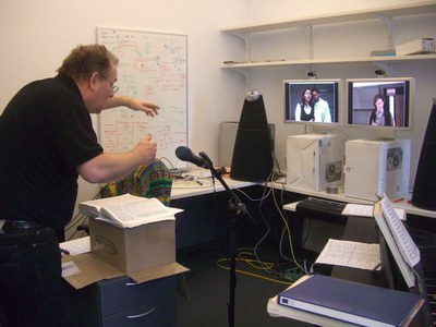 Niels Muus conducts in front of 2 screens showing three remote opera singers